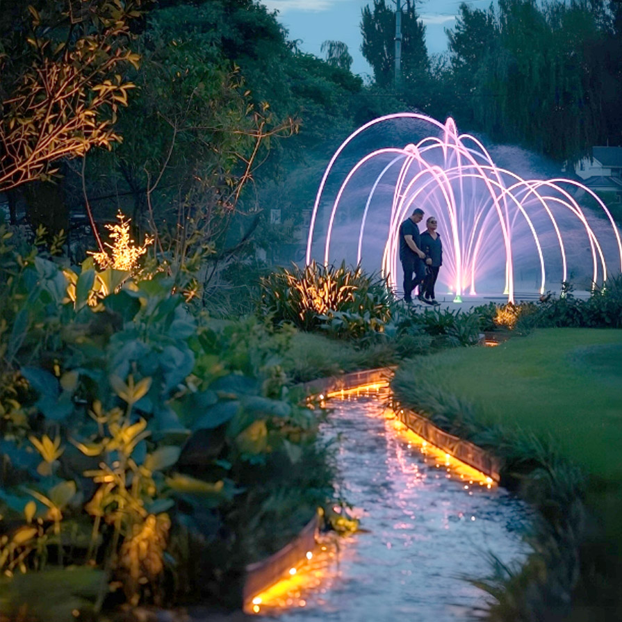 Ein Paar spaziert in einem Park. Ein leuchtender Bach und LED beleuchtete Bodensprinkler sind Teil der verträumten Landschaft.